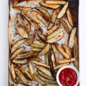 Cooked potato wedges on a parchment-lined sheet pan with a white dish of ketchup.