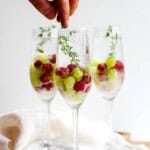 3 glasses with champagne mocktails in them with frozen grapes and someone placing a fresh thyme garnish inside the front glass.