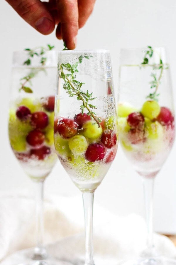 3 glasses with champagne mocktails in them with frozen grapes and someone placing a fresh thyme garnish inside the front glass.