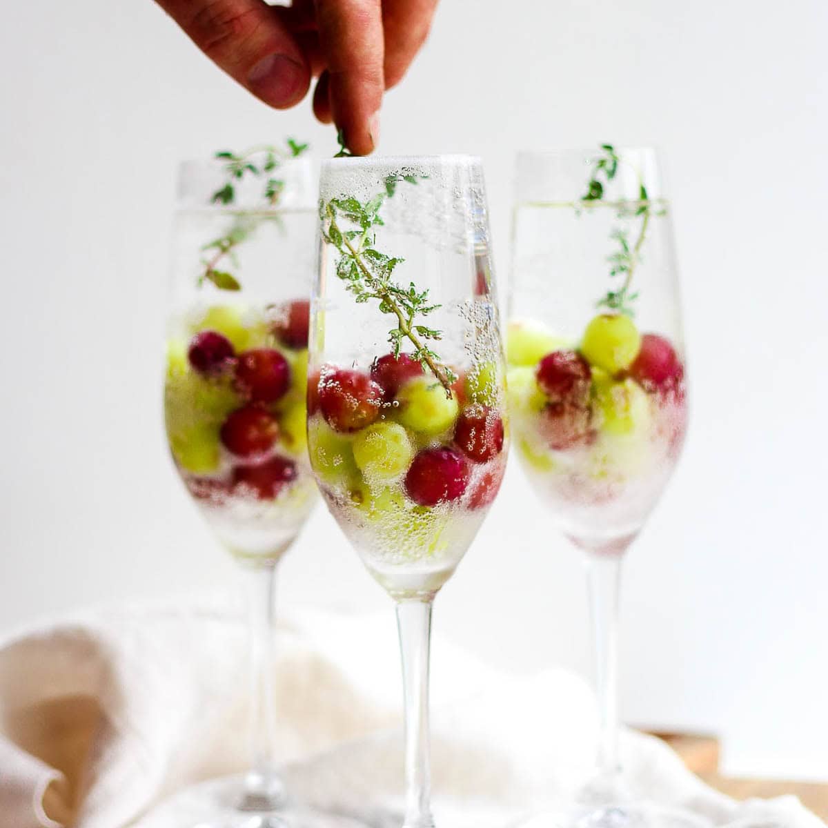 3 glasses with champagne mocktails in them with frozen grapes and someone placing a fresh thyme garnish inside the front glass.