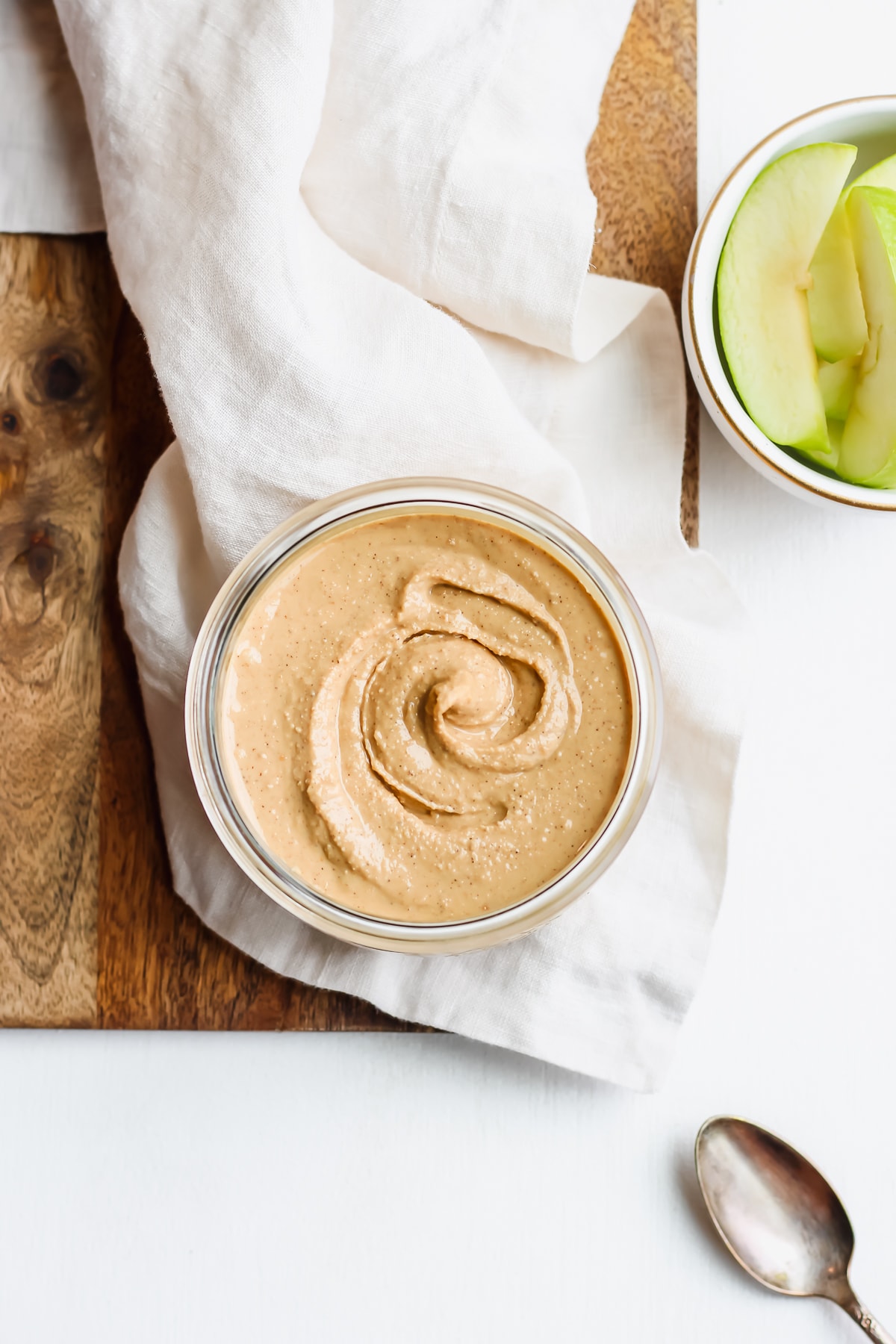 Jar of homemade cashew butter on a wooden board with a bowl of apples.