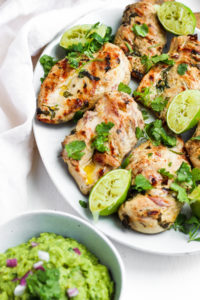 Grilled cilantro lime chicken breasts on a white platter with half limes and fresh cilantro next to a bowl of guacamole.