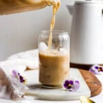 Someone pouring some bulletproof coffee into a cup with ice on a marble board with flowers next to it and coffee pot in the background.