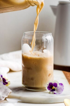 Someone pouring some bulletproof coffee into a cup with ice on a marble board with flowers next to it and coffee pot in the background.