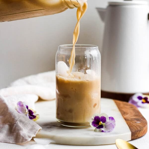 Someone pouring some bulletproof coffee into a cup with ice on a marble board with flowers next to it and coffee pot in the background.