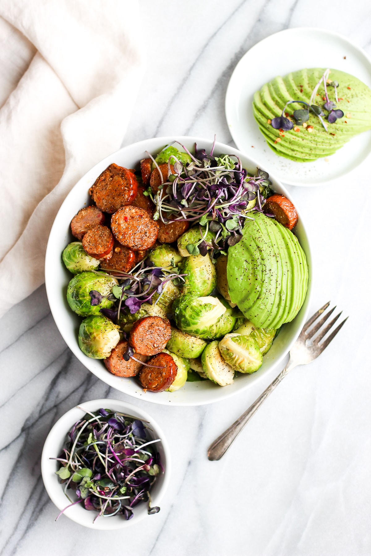 A white bowl with roasted chorizo and brussel sprouts, topped with avocado and micro-greens.