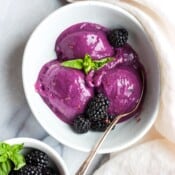 Three scoops of blackberry basil nice cream in a white bowl with fresh blackberries and a spoon sticking out the side.