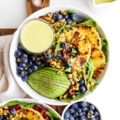 Seared pineapple in a bowl of salad with avocado, blueberries, and a small dish of dressing.