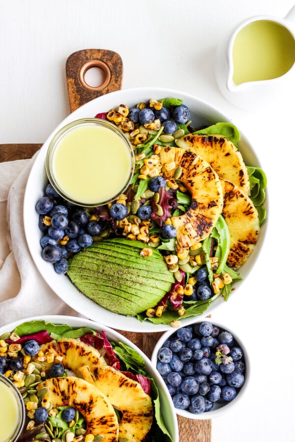 Seared pineapple in a bowl of salad with avocado, blueberries, and a small dish of dressing.