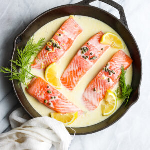 Four salmon fillets in a cast iron skillet with a creamy lemon caper sauce and fresh dill for garnish.