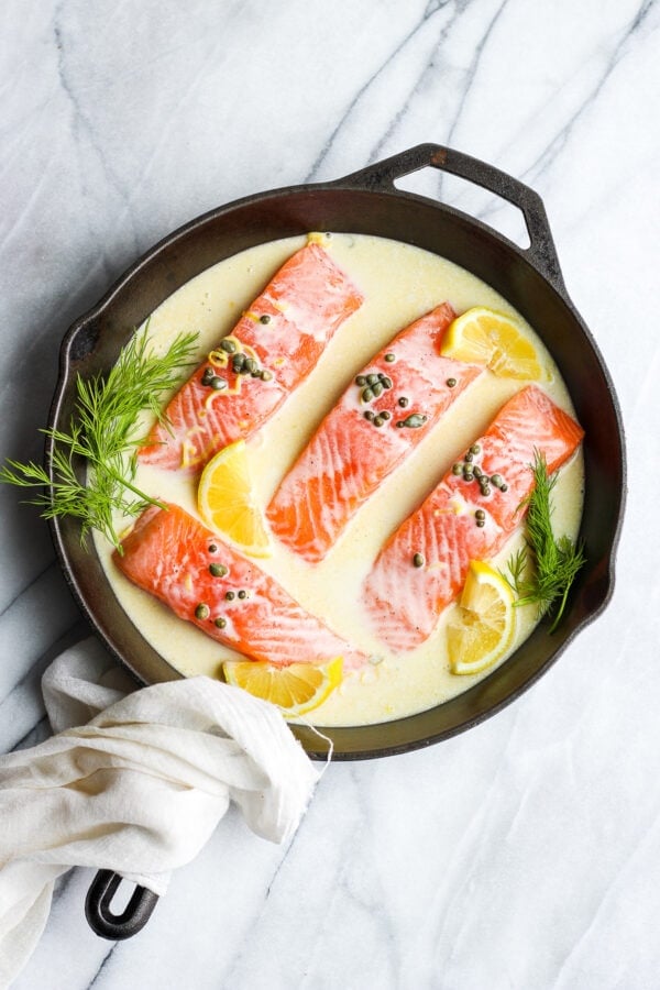 Four salmon fillets in a cast iron skillet with a creamy lemon caper sauce and fresh dill for garnish.
