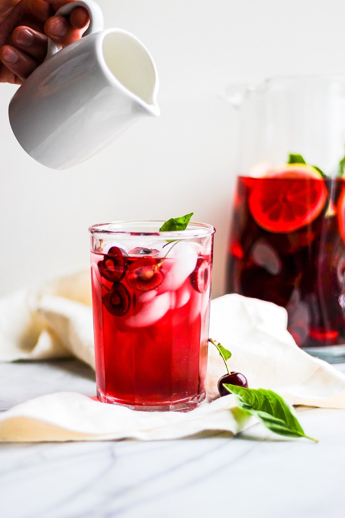 Glass of hibiscus tea on a marble board with someone pouring dairy-free creamer into it.  There is a pitcher of more tea in the background. 