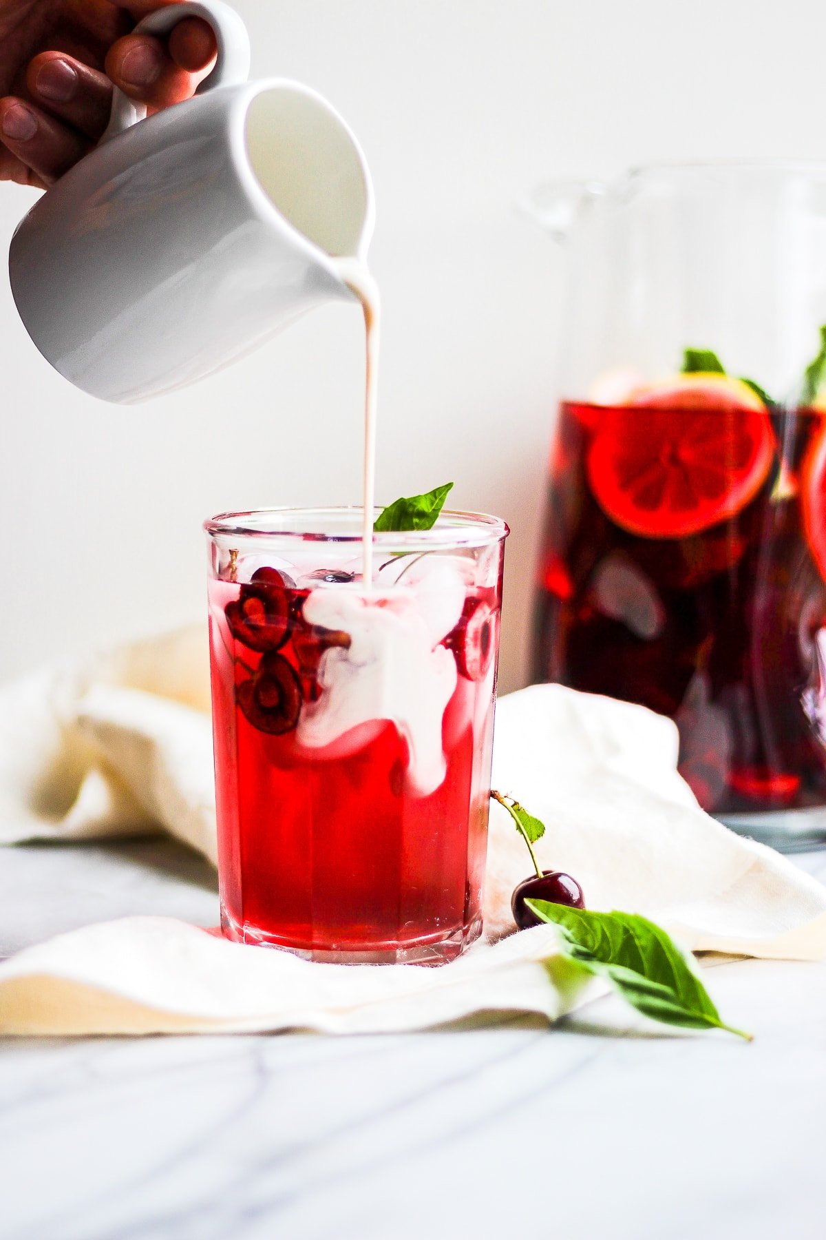 Glass of hibiscus tea on a marble board with someone pouring dairy-free creamer into it.  There is a pitcher of more tea in the background. 