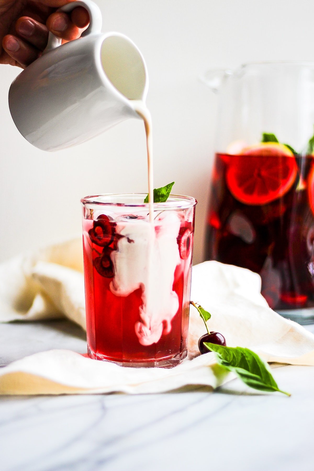 Glass of hibiscus tea on a marble board with someone pouring dairy-free creamer into it.  There is a pitcher of more tea in the background. 