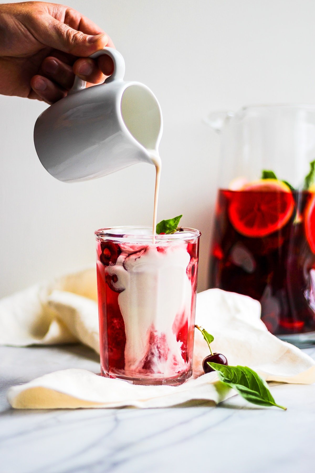 Glass of hibiscus tea on a marble board with someone pouring dairy-free creamer into it.  There is a pitcher of more tea in the background. 