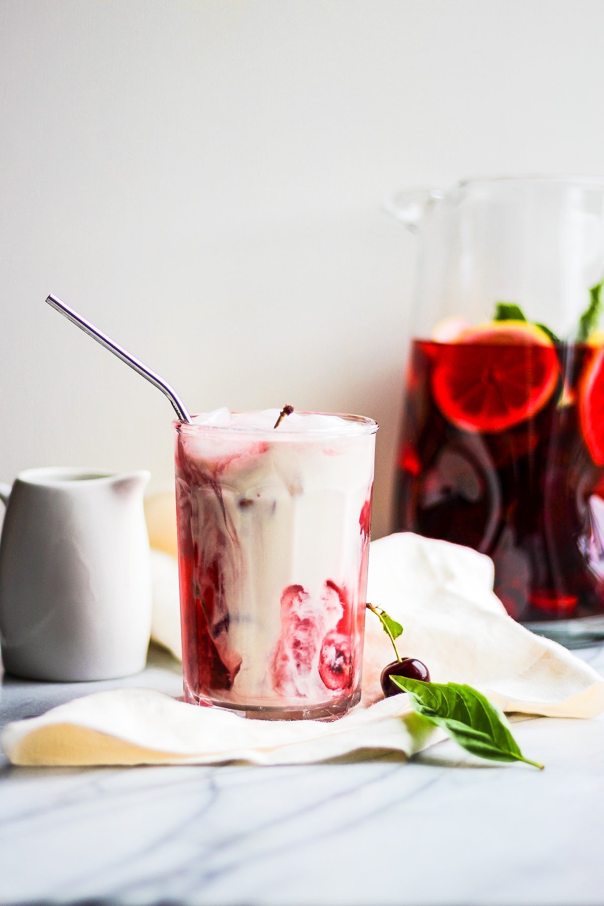 Glass of hibiscus tea on a marble board with dairy-free creamer in it.  There is a pitcher of more tea in the background. 