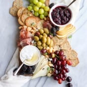 A cutting board full of gluten free crackers, deli meat, fresh fruit, olives, and two dishes of jam and cream & honey dip.