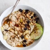 Apple pie oatmeal in a white bowl with a spoon and a sliced green apple on the side.
