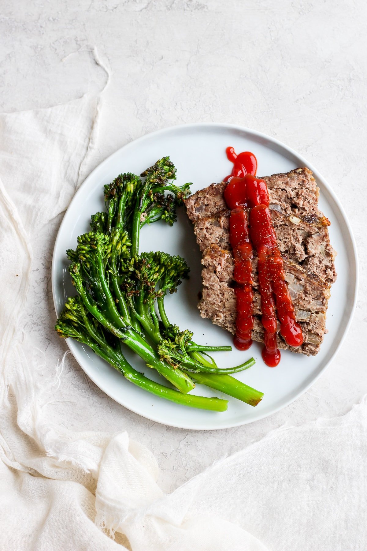 Sliced meatloaf on a plate with ketchup on top and roasted broccolini on the side.