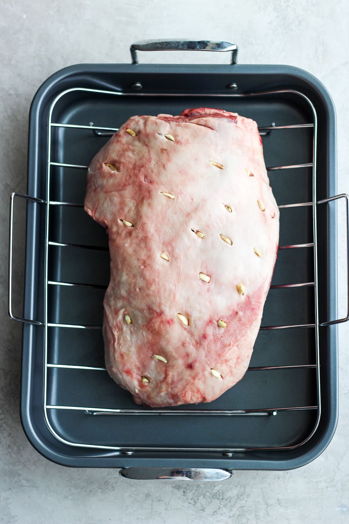 Roasted Rosemary Garlic Leg of Lamb sitting in a roasting pan with slits all over it and garlic slices sticking out. 