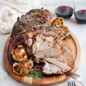A roasted leg of lamb on a cutting board with roasted heads of garlic on the side and glasses of red wine in the background.