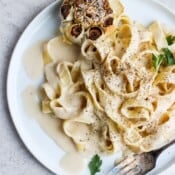 A white plate of cooked noodles with vegan alfredo sauce, a roasted head of garlic, and a fork.