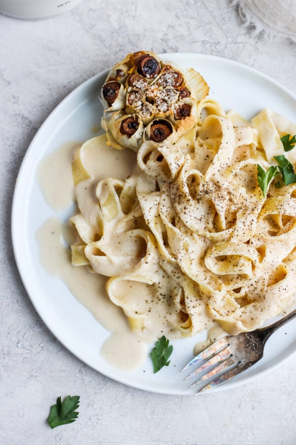 A white plate of cooked noodles with vegan alfredo sauce, a roasted head of garlic, and a fork.