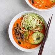 A bowl of Thai mushroom zoodle soup with chopsticks on the side of the bowl with another bowl of soup next to it.
