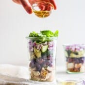 Two tall jars of greek chicken salad with a dish of oil being poured into one of them.