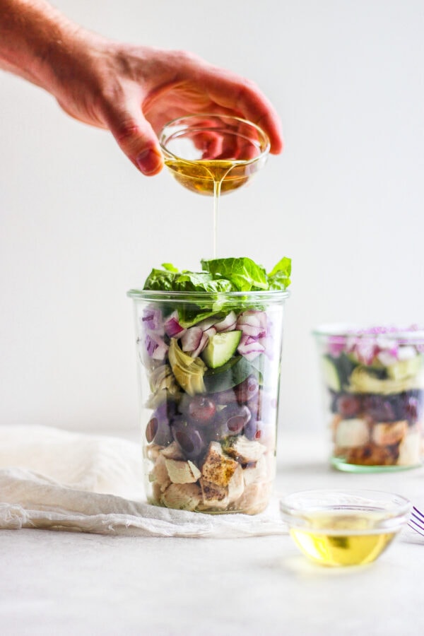 Two tall jars of greek chicken salad with a dish of oil being poured into one of them.