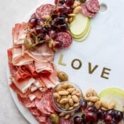 A large marble stone with the word "love" surrounded by deli meats, grapes, olives, nuts, and slices of green apple.