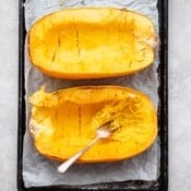 A fork starting to shred the inside of one of two halves of roasted spaghetti squash on a parchment-lined sheet pan.