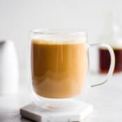 A glass mug of maple oat milk latte with a jar of syrup in the background.