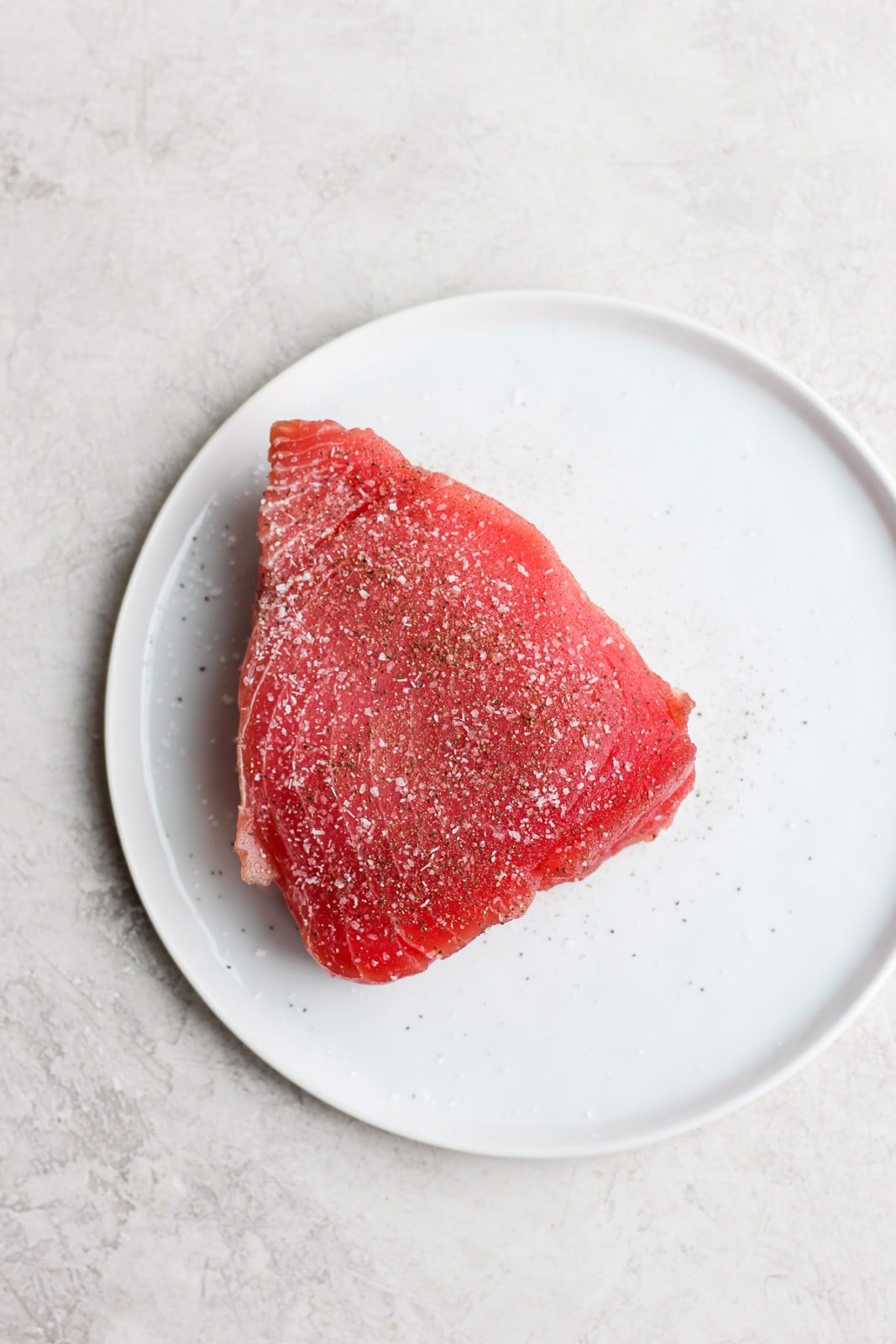 A piece of raw ahi tuna on a white plate sprinkled with salt and pepper. 