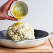 A head of cauliflower in a cast iron skillet with an olive oil & garlic mixture being poured on top.