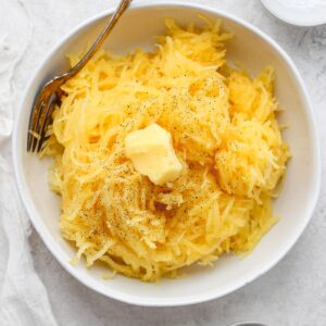 Shredded spaghetti squash in a white bowl with butter and a fork sticking out the side.