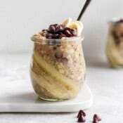 A small jar of chocolate chip banana overnight oats with a spoon, a couple chocolate chips in the foreground, and another jar of overnight oats in the background.