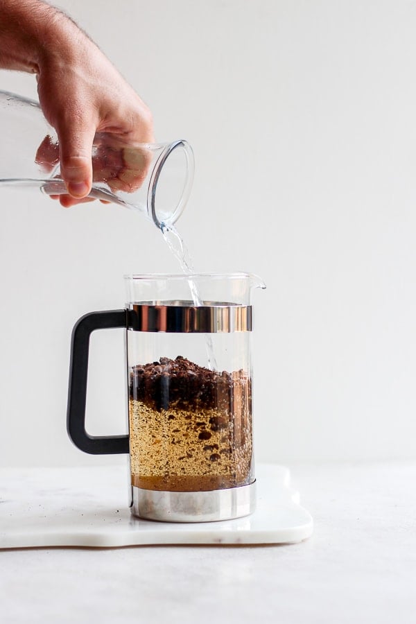 Someone pouring water into a glass french press which is mixing with roughly ground coffee beans.