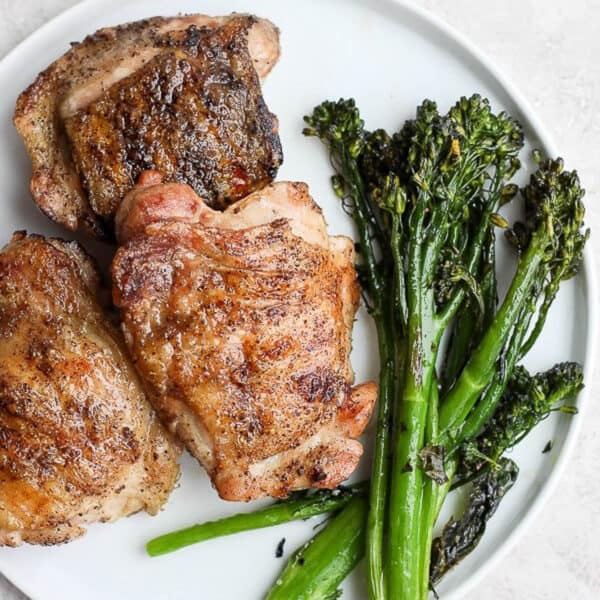 Three grilled chicken thighs on a plate next to some grilled broccolini.