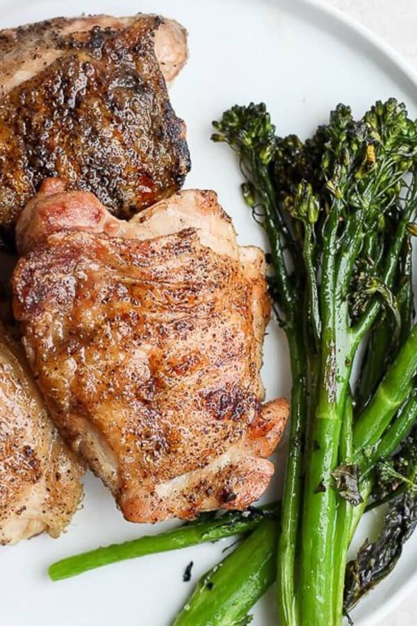Three grilled chicken thighs on a plate next to some grilled broccolini.
