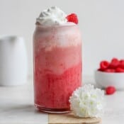 A raspberry smoothie in a tall glass with whipped cream and a small bowl of raspberries in the background.