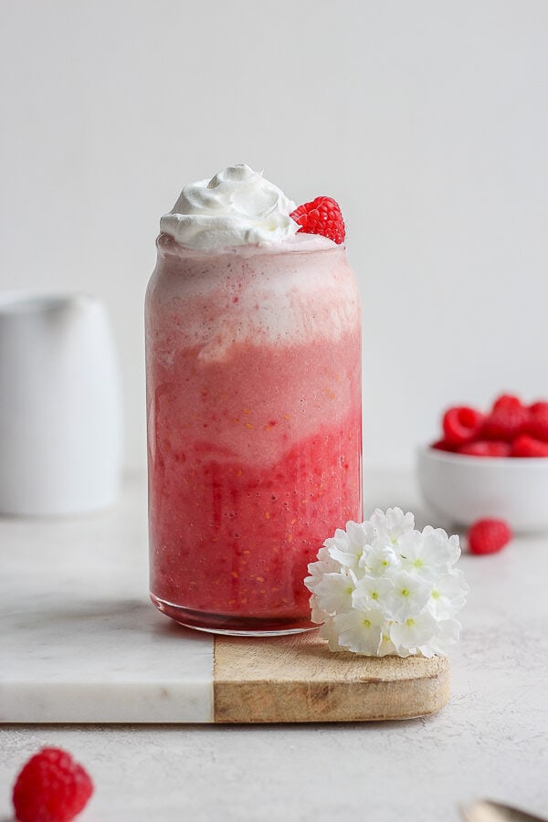 A raspberry smoothie in a tall glass with whipped cream and a small bowl of raspberries in the background.