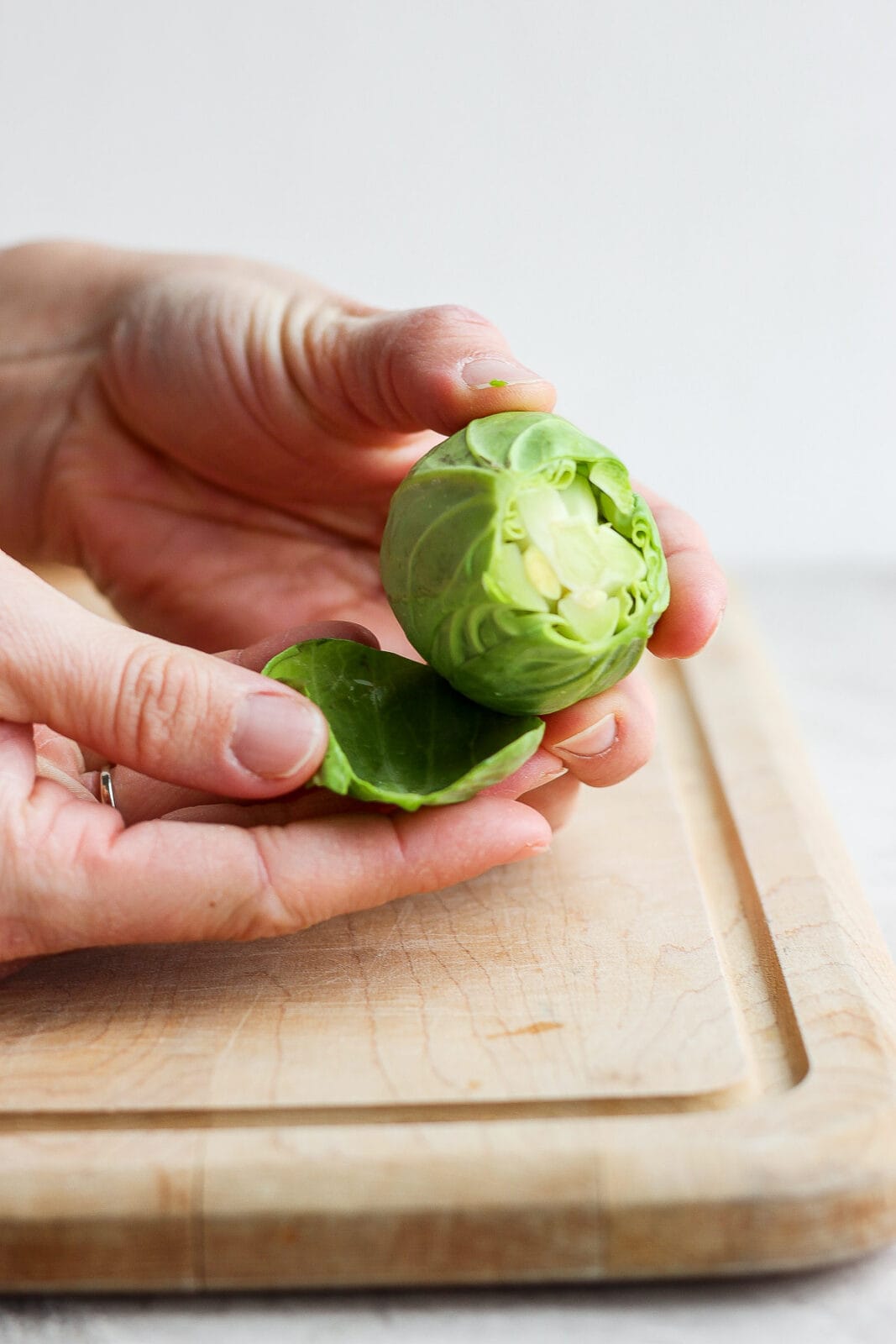 Someone holding a single brussel sprout with the end cut off and they are peeling back the outer layers to remove.