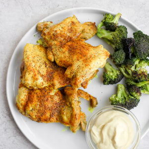 Stove top chicken thighs on a white plate with roasted broccoli and a small dish of dipping sauce.
