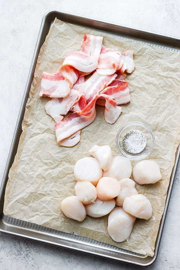 Scallops, bacon, and a dish of salt and pepper on a parchment-lined baking sheet.