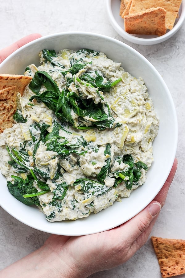 A bowl filled with vegan spinach artichoke dip with some crackers on the side. 