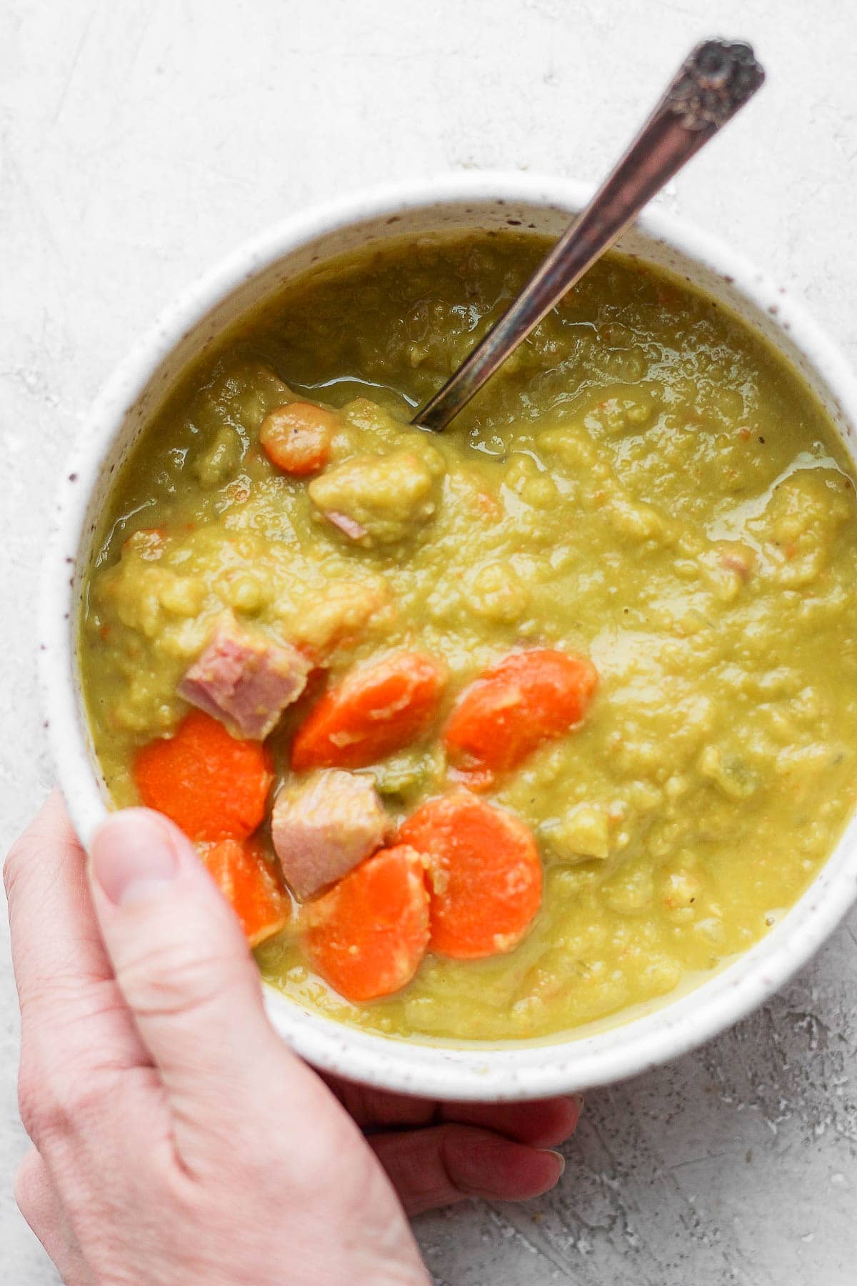 Bowl of split pea and ham soup with a spoon sticking out and a hand holding one side. 