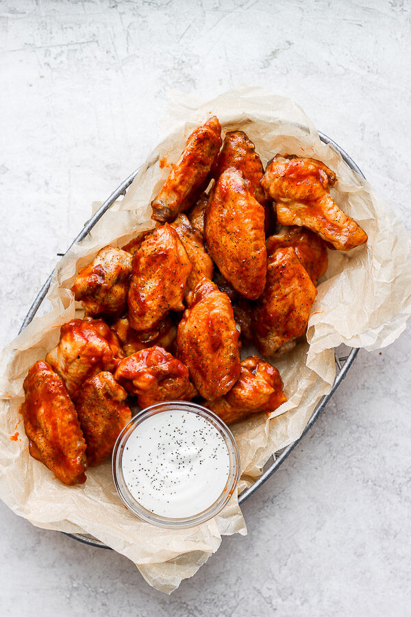 A metal basket full of bbq chicken wings and a small dish of ranch dressing.