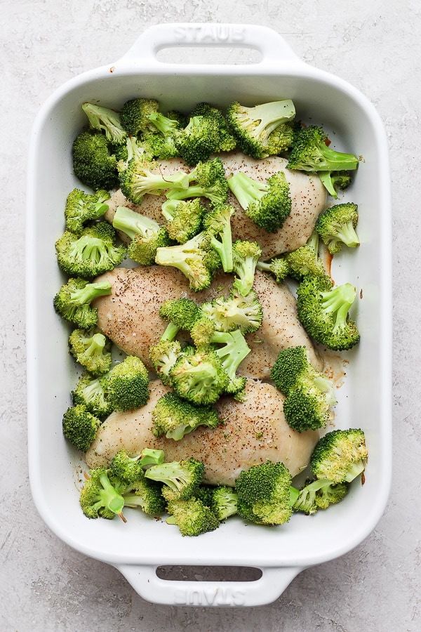 White baking dish with chicken partially cooked and broccoli added.