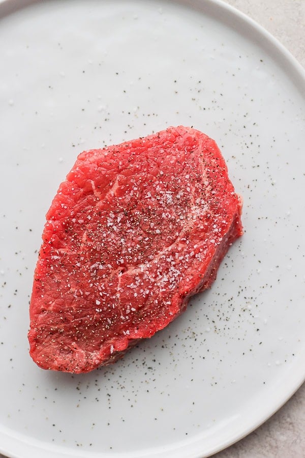 A raw seasoned steak on a white plate.